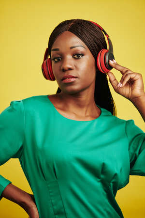 Vertical waist up studio portrait of stylish young Black woman listening to music in her headphones looking at cameraの写真素材