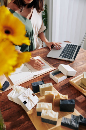 Woman planning strategy of selling handmade soap via online shop and taking notes in textbook