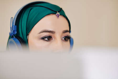Young beautiful software engineer in hijab listening to music in headphones when programming or editing video on computerの写真素材