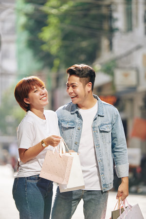 Smiling young Vietnamese man giving shopping-bags with presents to his happy excited young girlfriendの写真素材