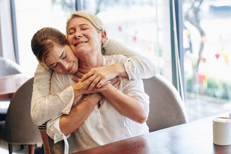 Happy beautiful young woman hugging her mature mother from behind when they are spending time in cafeの写真素材