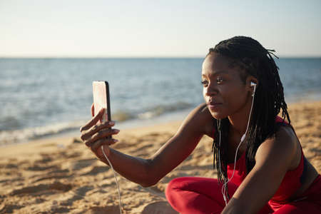 Positive pretty young sportswoman resting on beach after jogging and filming video for her subscribersの写真素材