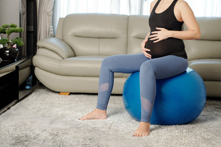 Cropped image of slim pregnant woman touching her belly when resting on yoga ball after working out