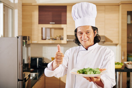 Smiling mature Asian chef holding plate with dish he cooked and showing thumbs-up