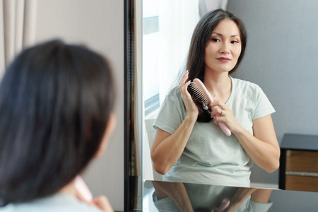 Beautiful young Vietnames woman sitting in front of mirror and enjoying brushing hairの写真素材