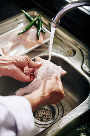 Hands of senior person rinsing chicken thights under cold water from kitchen tapの写真素材