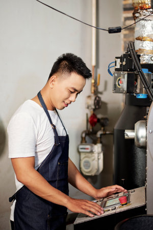 Smiling Vietnamese young man wearing denim apron when operating new coffee roasting machine