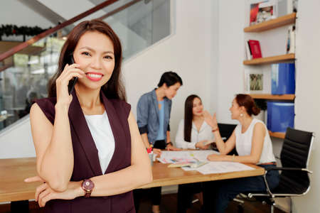 Beautiful happy young female entrepreneur talking on phone and looking at cameraの写真素材