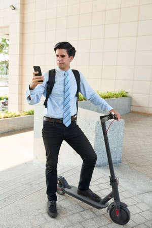 Young businessman with electric kick scooter checking text messages in his smartphoneの写真素材