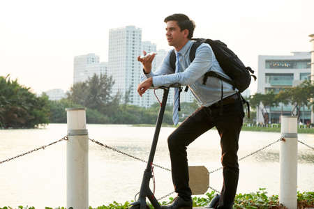 Young businessman standing outdoors with electric scooter and recording voice message for coworkerの写真素材