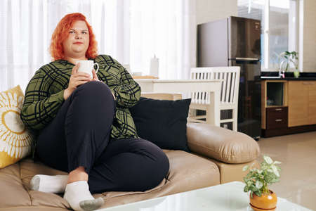 Pensive red haired woman in plaid shirt resting on sofa with big cup of coffeeの写真素材