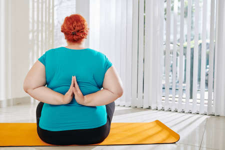 Rear view of overweight woman sitting on yoga mat and keeping hands in reverse namaste mudra behind her backの写真素材
