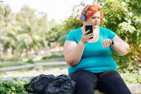 Overweight young woman checking data in her fitness tracker after exercising in city parkの写真素材