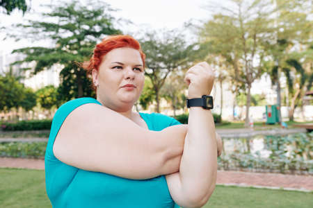Serious plus size young woman doing stretching exercise for her arms when warming up in parkの写真素材
