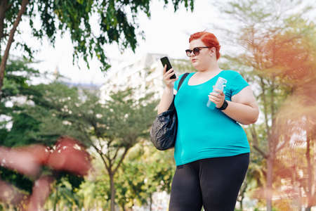 Overweight pretty young woman standing in city park and checking health application on smatphone after trainingの写真素材