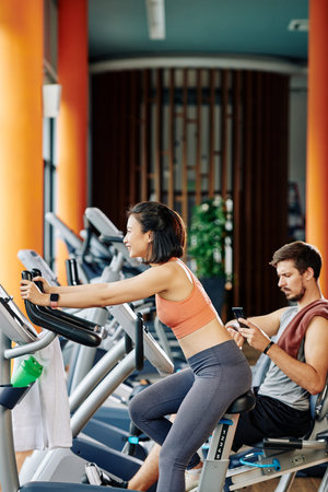 Smiling pretty young Asian woman training at fitness center and riding stationary bike