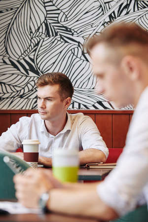 Serious frowning young businessman working on laptop at local cafe and drinking cup of coffeeの写真素材