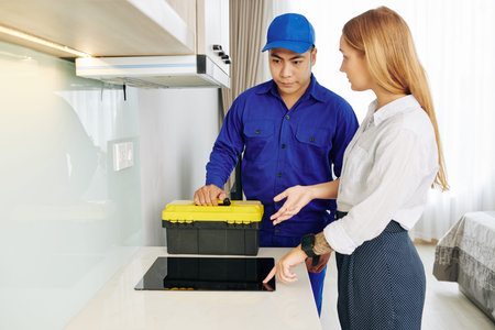 Pretty young woman asking electrician with toolbox to check her broken induction cooker