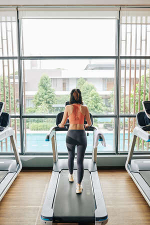 Slim young woman walking on treadmill in gym with big panoramic windowsの写真素材