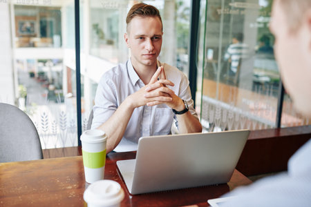 Smiling young businessman sitting at cafe table with opened laptop and interviewing job applicant