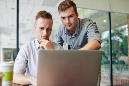 Young entrepreneur helping colleague with finishing project for report, he is pointing at laptop...