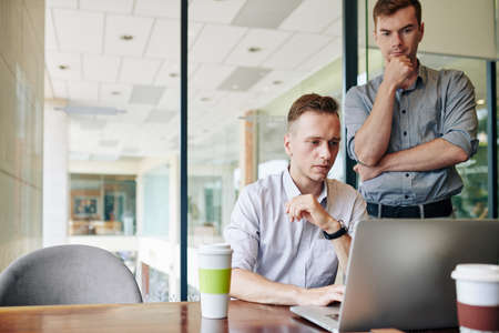 Pensive software developer looking at his colleague coding or making presentation on laptop at office deskの写真素材