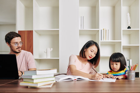 Software developer working on laptop at home when wife teaching their daughter writing at the same tableの写真素材