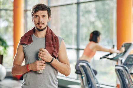 Portrait of handsome fit young man with towel on shoulder drinking fresh water after training in gymの写真素材