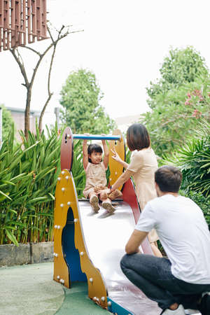 Happy young parents helping their daughter to slide down at playgroundの写真素材