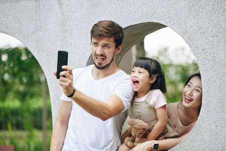 Young man making funny face when photographing with his wife and little daughter in city parkの写真素材