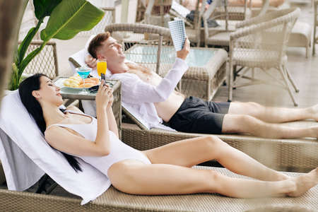 Beautiful happy young couple resting in chaise-lounges by swiming pool, reading book and checking social mediaの写真素材