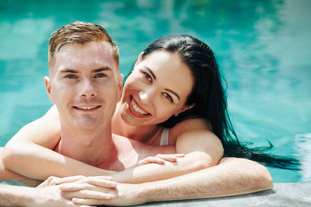 Beautiful young smiling woman hugging boyfriend from behind when they are spending time in swimming...