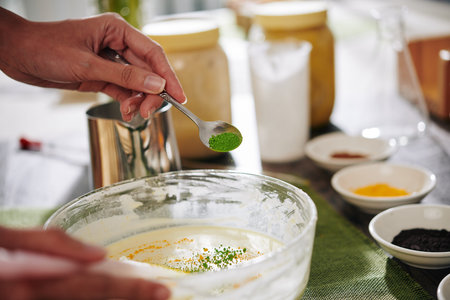 Creative woman putting spoon of green and yellow powder colorants in soap mixture