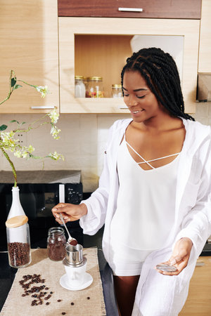 Smiling woman in sleepwear putting spoon of ground coffee brewing pot when making Vietnamese style...