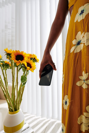 Cropped image of young woman with smartphone in hand standing next to coffee table with vase of...