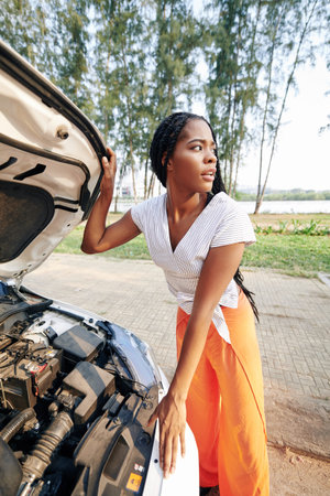 Stressed woman opening hood of broken car and looking around searching for help