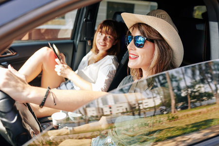Happy young woman in straw hat and sunglasses driving car and talking to her friend
