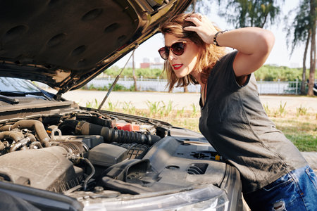 Stressed young woman opening car hood and looking at engine of broken carの写真素材