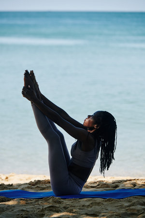 Young fit Black woman in seated balancing yoga pose exercising on beach