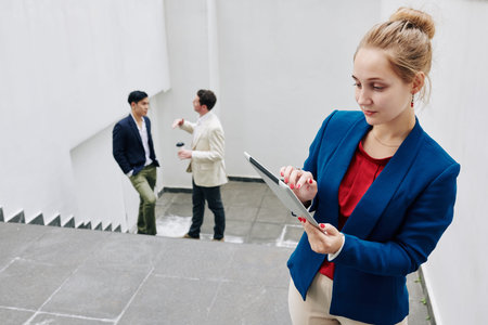 Young female entrepreneur reading article on tablet computer, her colleagues talking in background