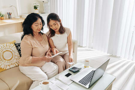 Young Asian woman helping her senior mother to figure out how to pay bill online via computerの写真素材