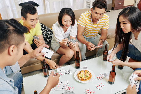 Group of cheerful friends drinking beer and eating pizza when playing cards at house party