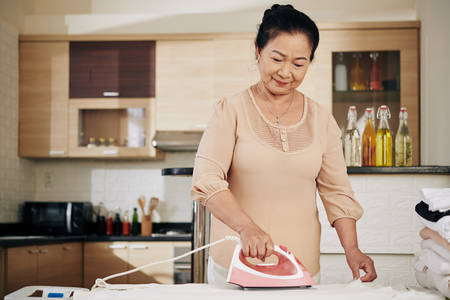 Smiling senior Asian woman ironing clothes at home during pandemic period when she needs to stay at homeの写真素材