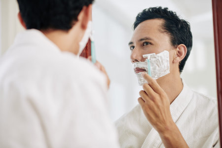 Serious mixed-race man looking at bathroom mirror and shaving off his beard