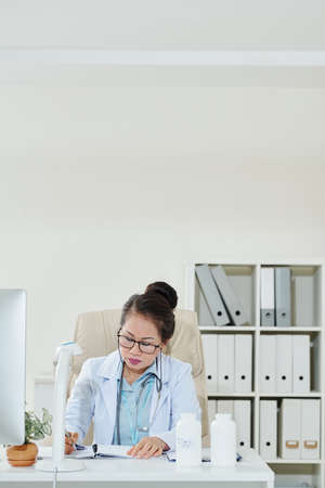 Serious mature female doctor sitting at office desk and taking notes in plannerの写真素材