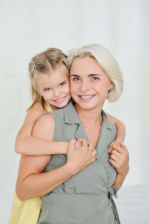 Portrait of happy smiling pretty blond woman and little daughter hugging her from behindの写真素材
