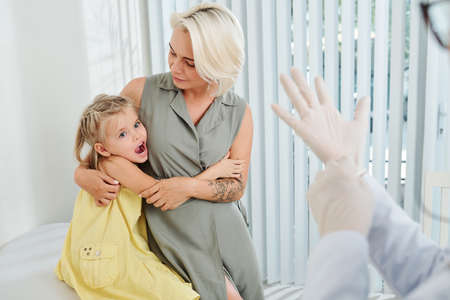 Scary little girl hugging her mother and looking at doctor putting on rubber glovesの写真素材