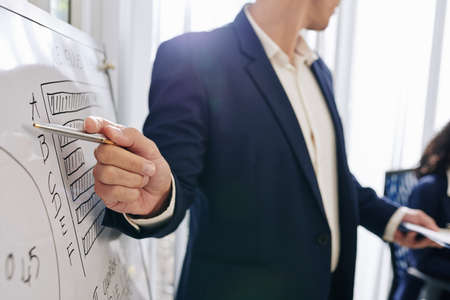 Close-up image of entrepreneur pointing at diagram on whiteboard when talking in front of colleagues about company developmentの写真素材
