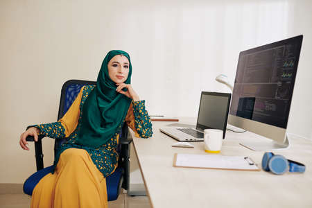 Confident muslim female software developer in traditional dress in hijab sitting at her office tableの写真素材
