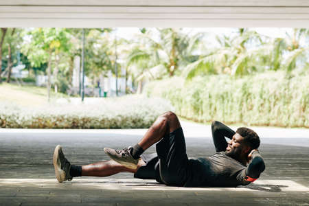 Happy young Black sportsman lying on ground and doing bicycle crunches to improve his absの写真素材